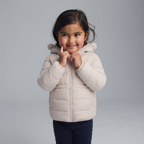 Niña sonriente vistiendo chaqueta acolchada beige con capucha de osito de la marca EmpiBy