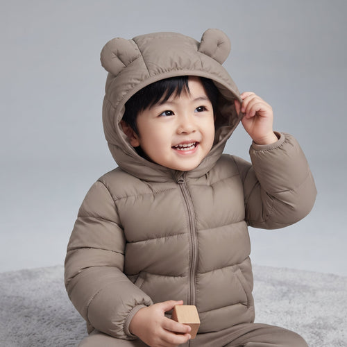 Bebé sonriente vistiendo chaqueta acolchada EmpiBy color topo con capucha y orejitas de oso sobre fondo gris