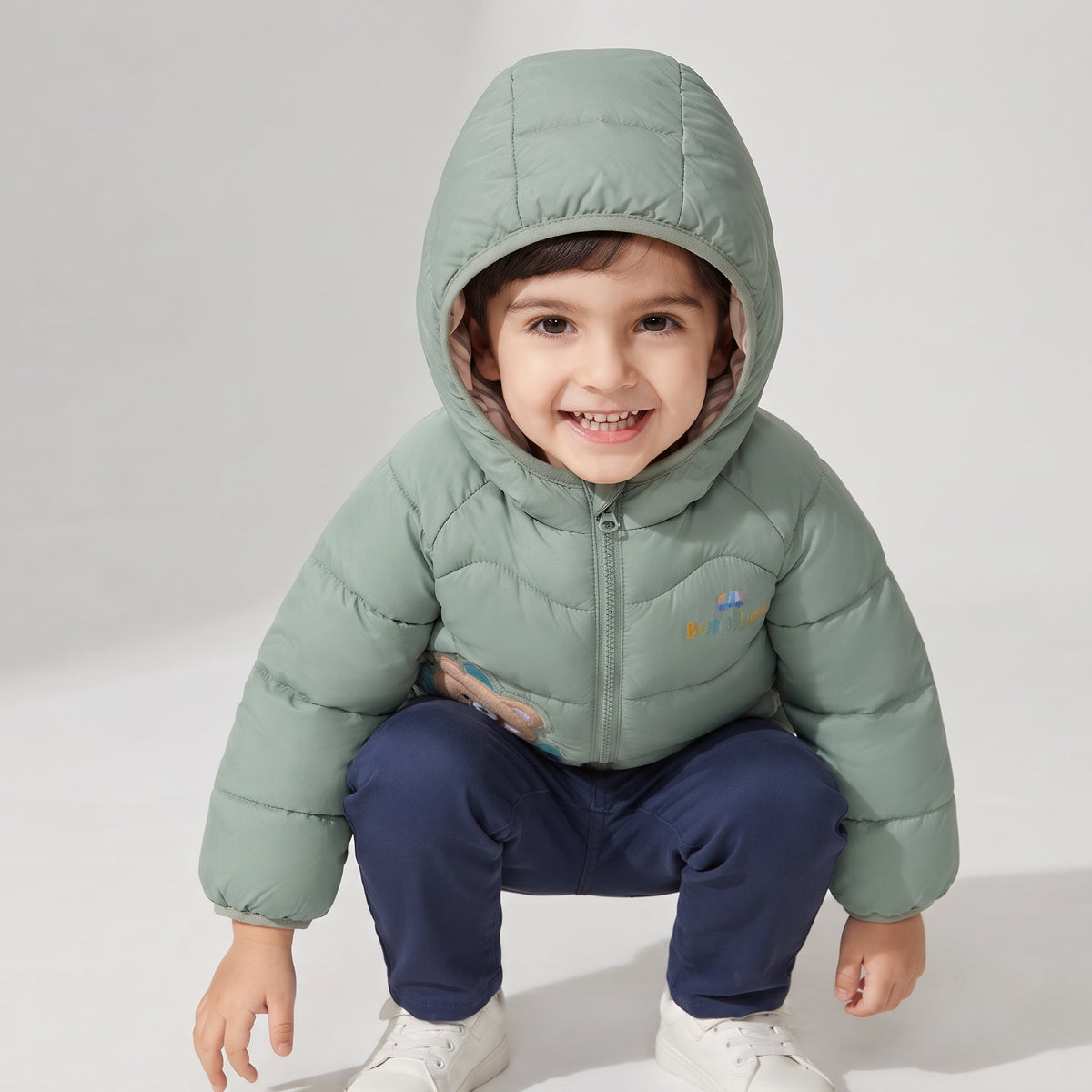 Niño sonriente con chaqueta acolchada verde menta con capucha y diseño de osito de la marca EmpiBy