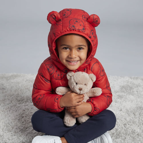 Niño sonriente con chaqueta acolchada roja EmpiBy con estampado de osos y capucha con orejas, abrazando un peluche.