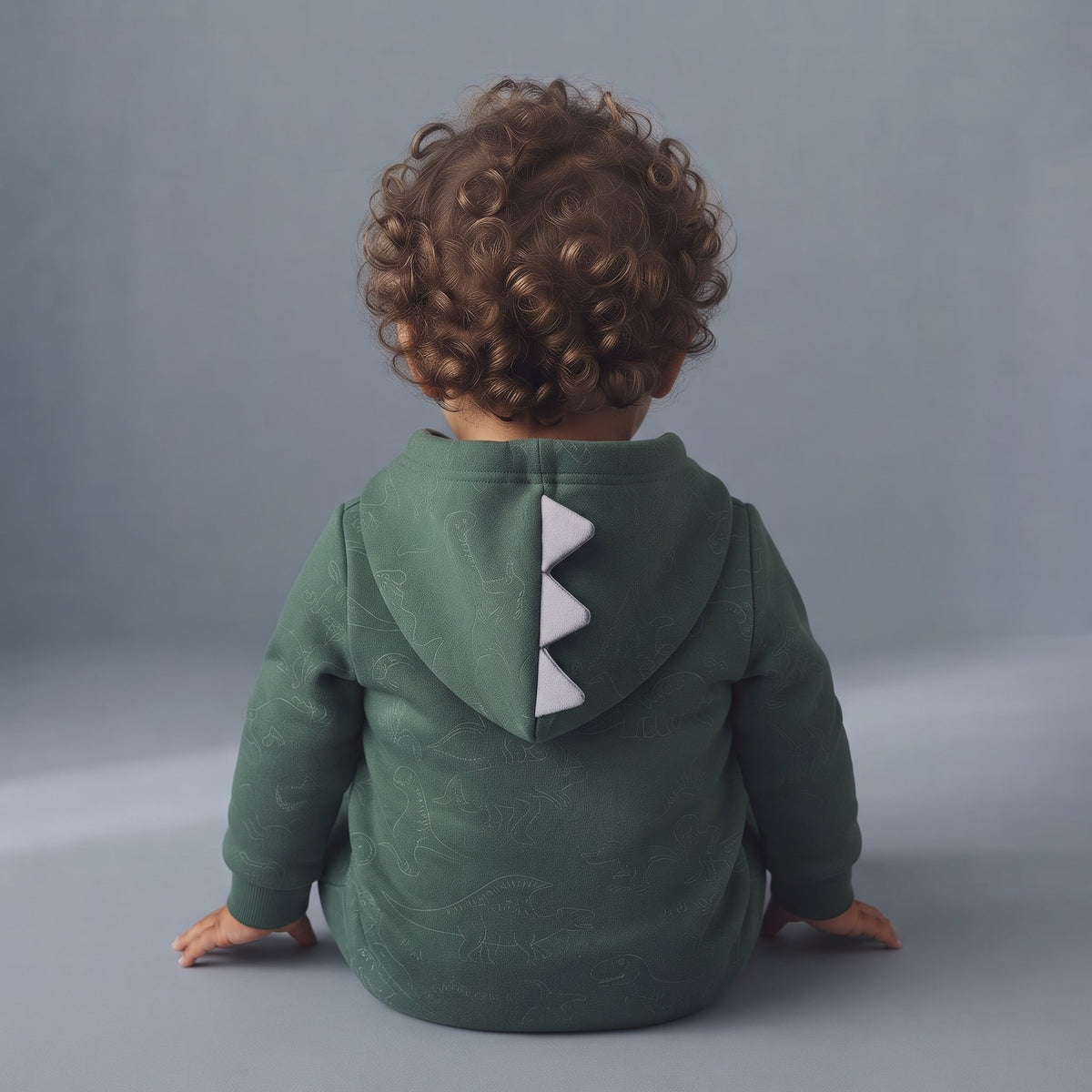Bebé de espaldas con chaqueta verde EmpiBy con capucha y cresta de dinosaurio gris, estampado de dinosaurios sutil.
