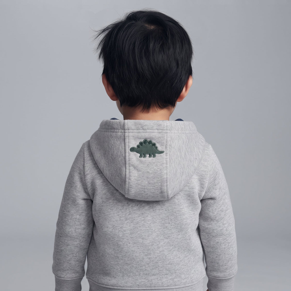 Niño de espaldas vistiendo una chaqueta gris con capucha de EmpiBy que tiene un dinosaurio bordado en la parte trasera