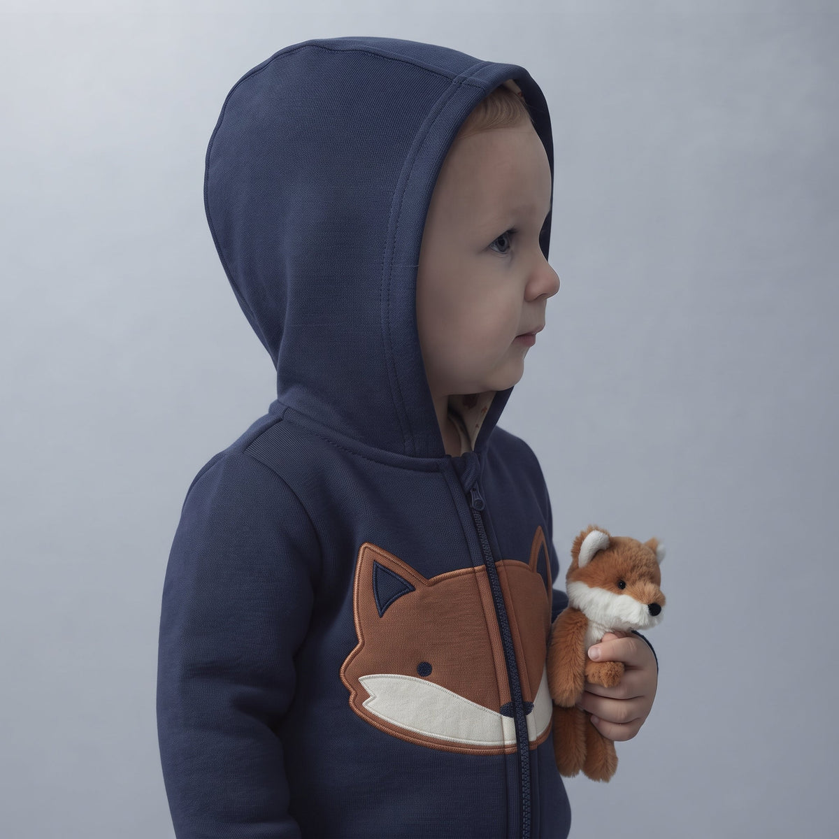 Niño pequeño con chaqueta azul marino con capucha y diseño de zorro de EmpiBy, sosteniendo un peluche de zorro.