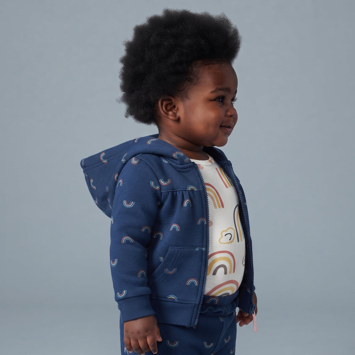 Bebé con conjunto EmpiBy de 3 piezas: chaqueta azul con capucha, body blanco y pantalón, todos con estampado arcoíris.