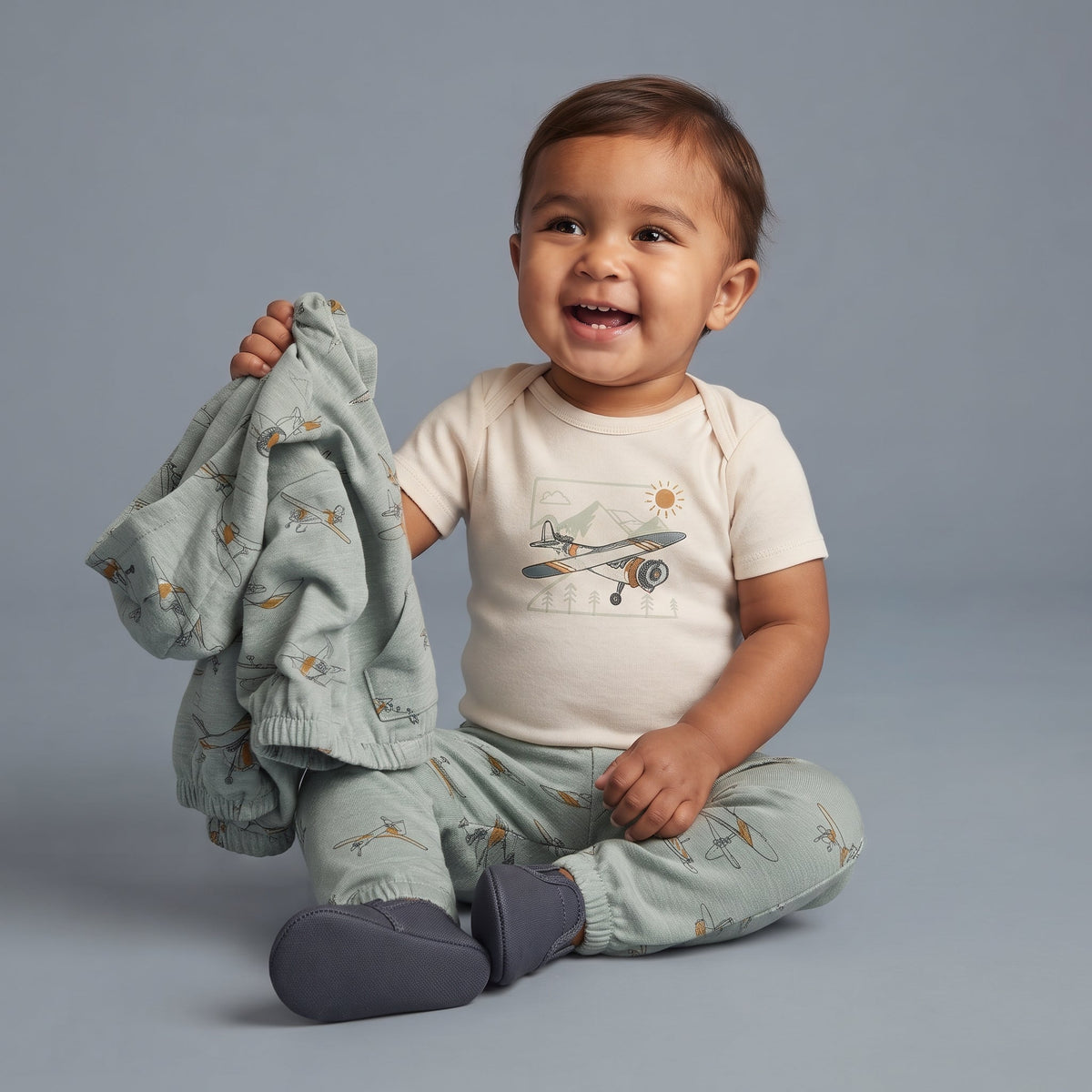 Bebé sonriente con conjunto EmpiBy de 3 piezas: body beige, pantalón y chaqueta verde salvia con estampado de aviones.