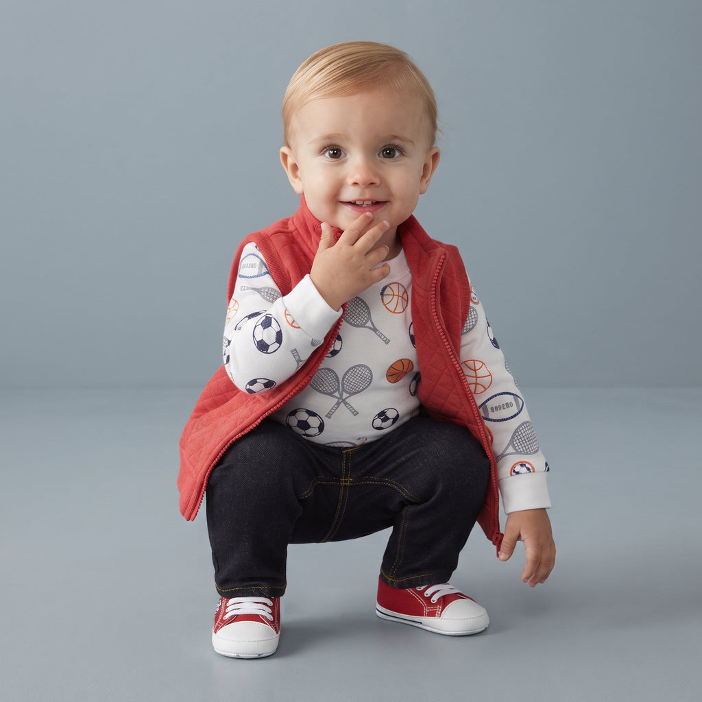 Bebé sonriente con conjunto EmpiBy de 3 piezas: chaleco rojo acolchado, body estampado y pantalón oscuro de mezclilla.