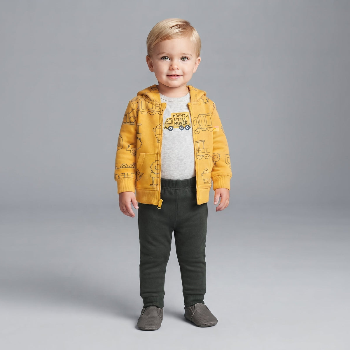 Niño pequeño vistiendo conjunto EmpiBy de 3 piezas: chaqueta amarilla con camiones, body gris y pantalón oscuro.