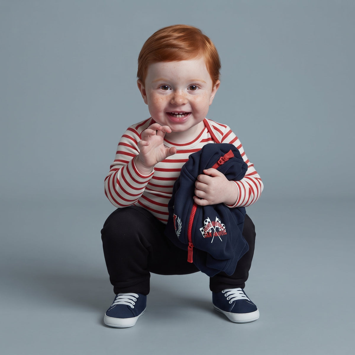 Bebé pelirrojo con conjunto EmpiBy de body a rayas, chaqueta azul con parches y pantalón negro sobre fondo gris.