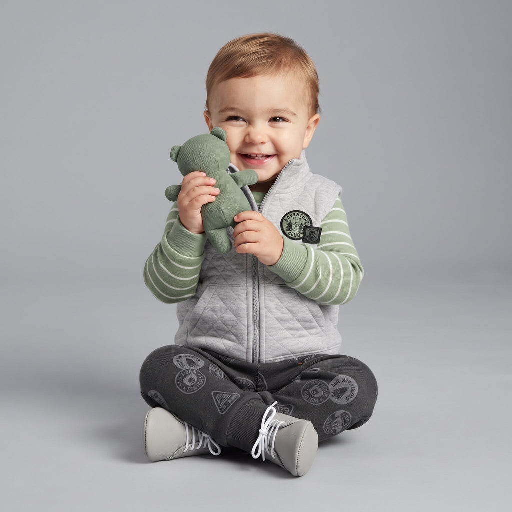 Bebé sonriente con conjunto EmpiBy de 3 piezas: chaleco gris acolchado, body a rayas verde y pantalón estampado.
