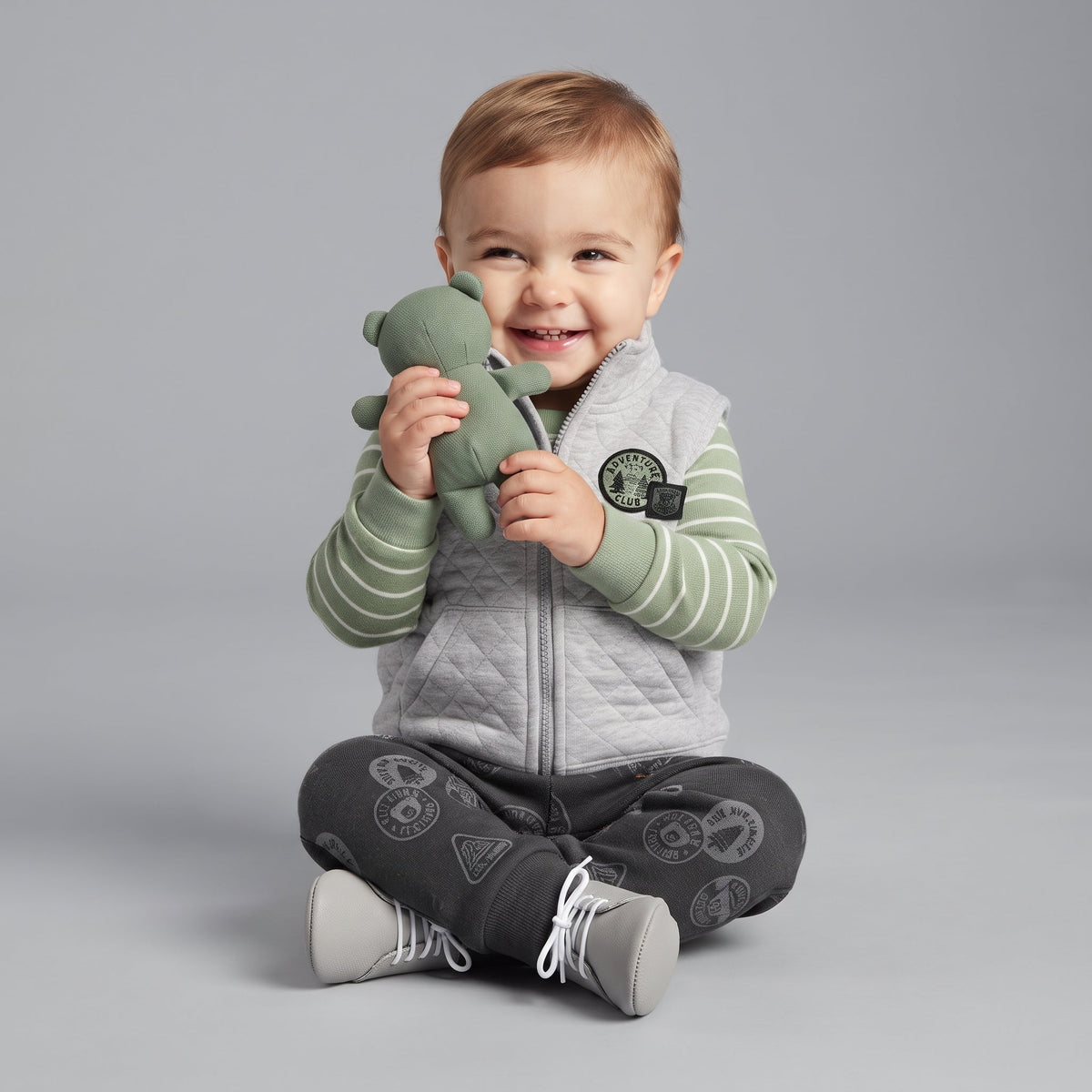 Bebé sonriente con conjunto EmpiBy de 3 piezas: chaleco gris acolchado, body a rayas verde y pantalón estampado.