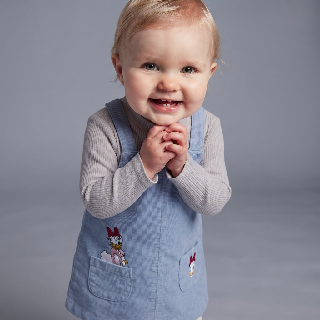Bebé sonriente vistiendo conjunto EmpiBy de 3 piezas con vestido tipo peto azul de Daisy Duck y camiseta gris