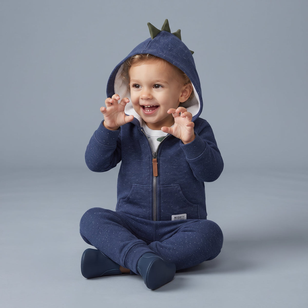 Bebé niño sonriente con conjunto EmpiBy de 3 piezas azul, chaqueta con capucha de dinosaurio y body estampado.