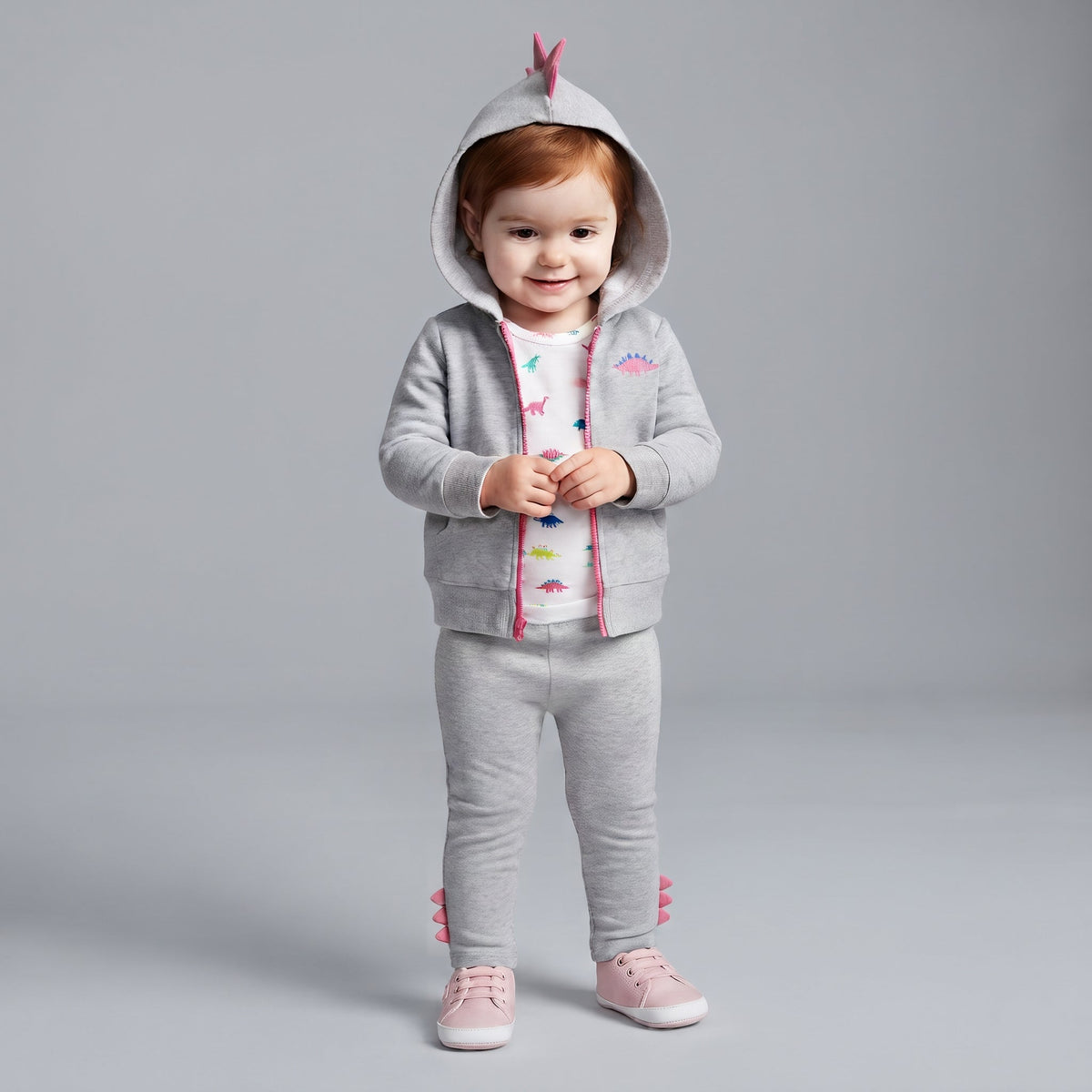 Bebé niña con conjunto EmpiBy de 3 piezas: chaqueta gris con capucha de dinosaurio, body estampado y pantalón a juego.