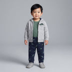 Niño pequeño vistiendo conjunto EmpiBy de 3 piezas: chaqueta gris, body a rayas verde y pantalón azul con dinosaurios.