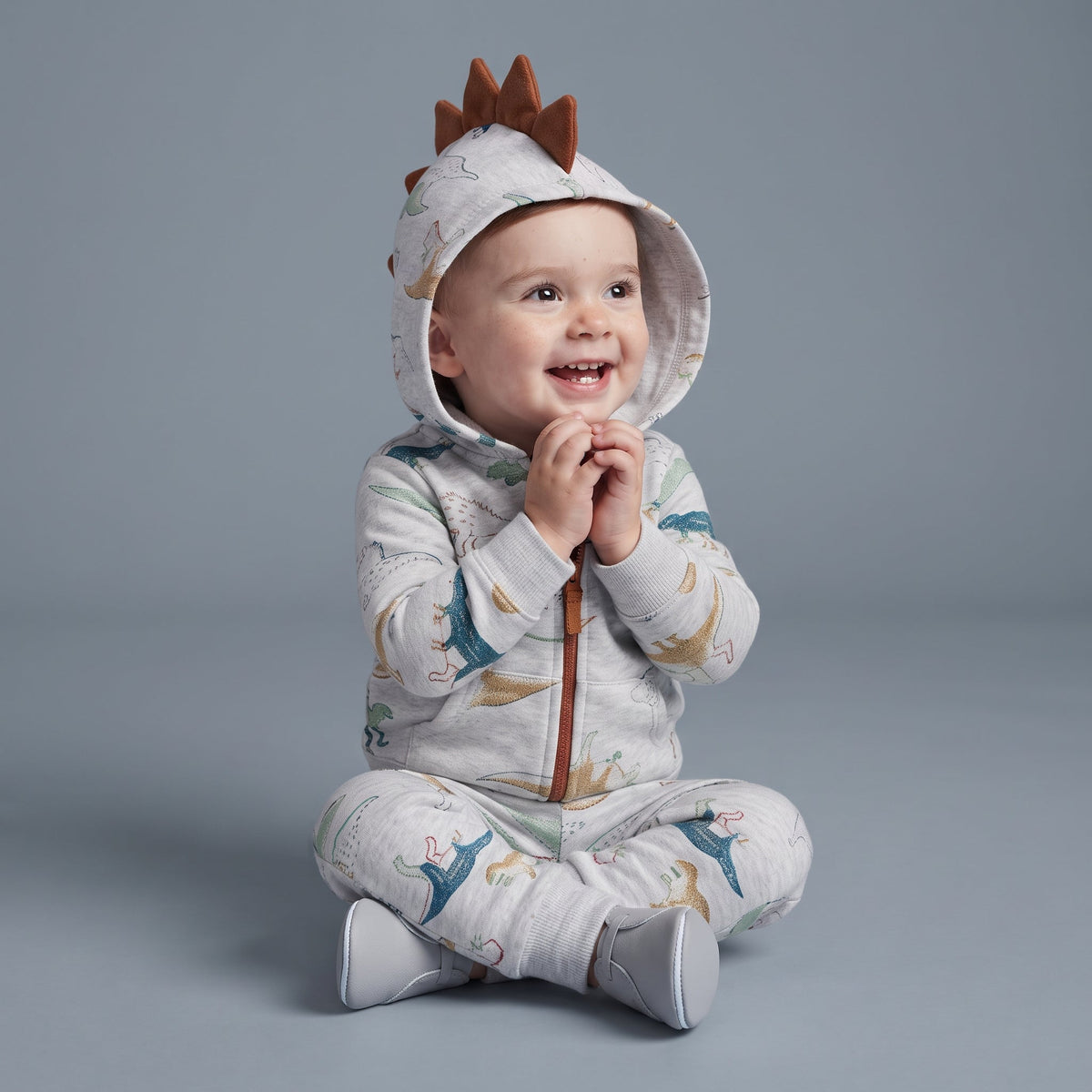 Bebé sonriente con conjunto EmpiBy de 3 piezas gris con estampado de dinosaurios y capucha con cresta de dinosaurio