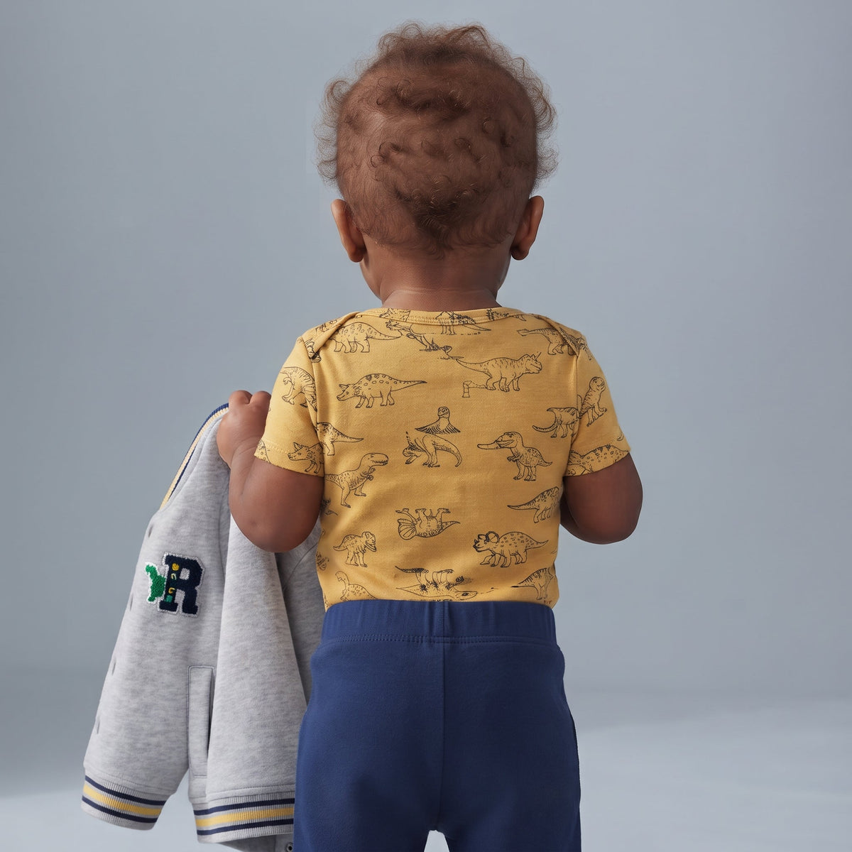 Bebé de espaldas vistiendo conjunto EmpiBy con body amarillo de dinosaurios, pantalón azul y chaqueta gris en mano