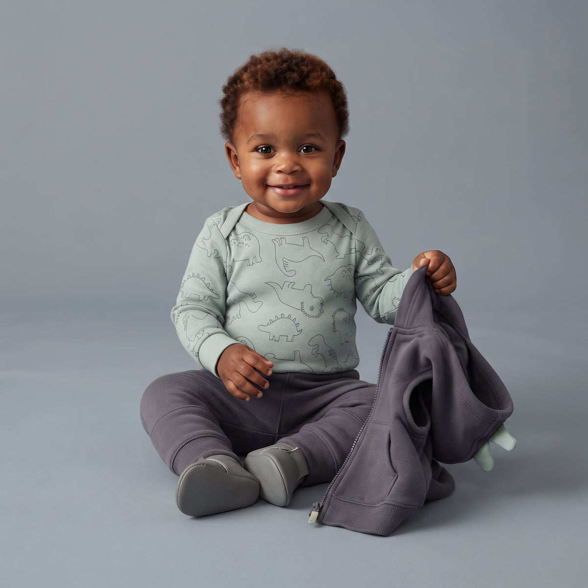 Bebé sonriente con conjunto EmpiBy de body verde con dinosaurios, pantalón gris y chaleco con capucha de picos