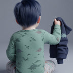 Bebé con body verde oliva de dinosaurios EmpiBy, sosteniendo una chaqueta azul con estampado de estegosaurio