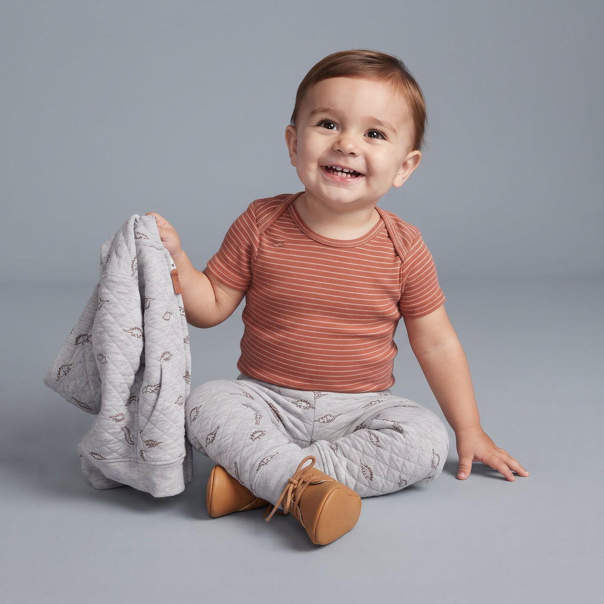 Bebé sonriente con conjunto EmpiBy de 3 piezas: body terracota a rayas, pantalón y chaqueta acolchada de dinosaurios.
