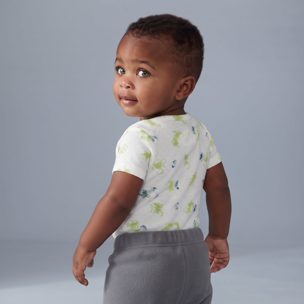 Bebé con body blanco estampado de dragones verdes y pantalón gris de EmpiBy, vista trasera sobre fondo neutro.