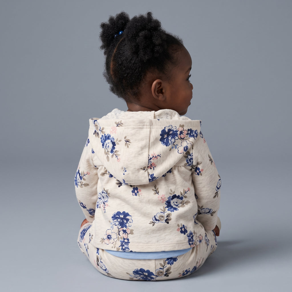 Bebé niña de espaldas vistiendo conjunto EmpiBy de chaqueta y pantalón con estampado floral azul sobre fondo gris.