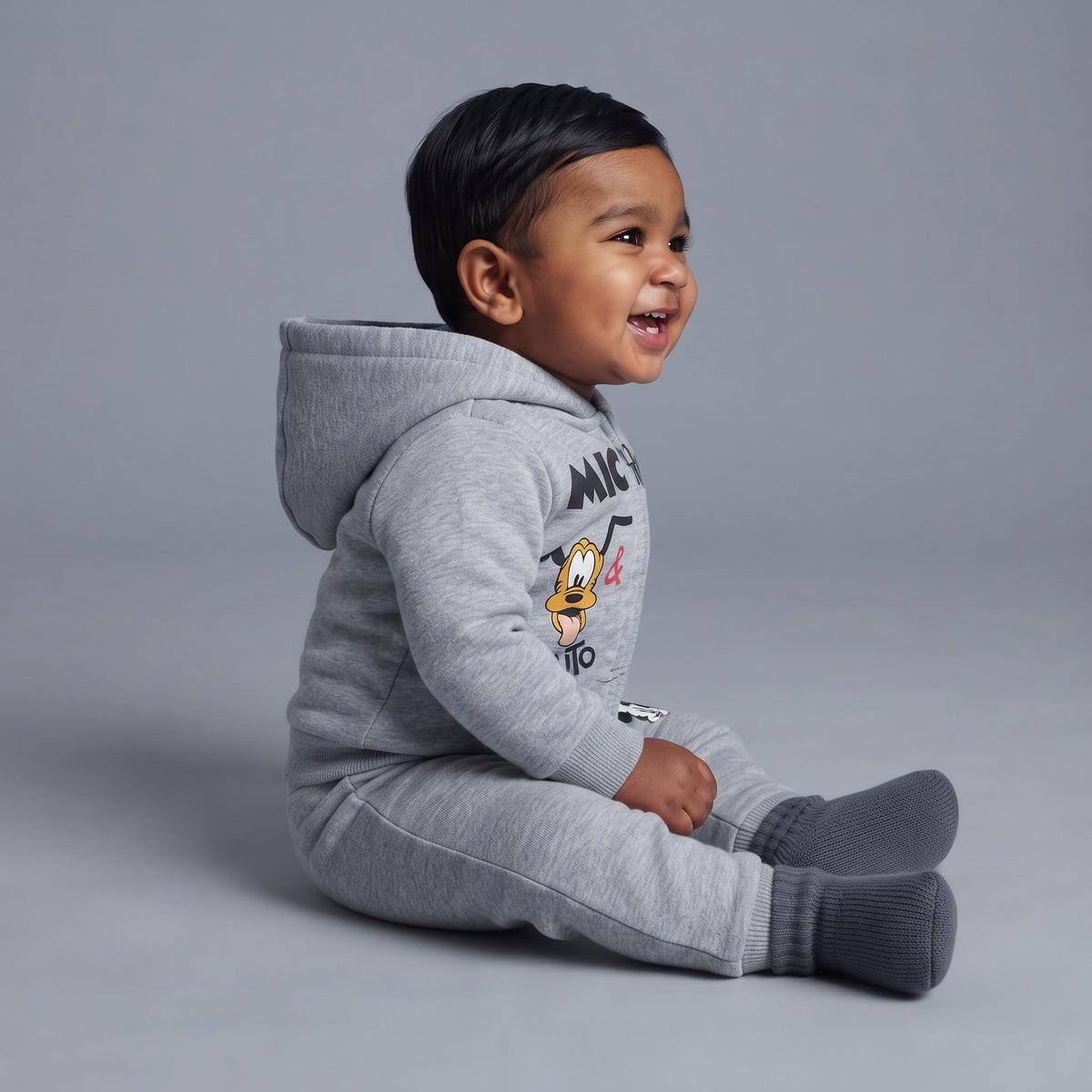 Bebé sonriente sentado con conjunto EmpiBy de dos piezas gris jaspeado de Mickey Mouse y Pluto con sudadera y jogger