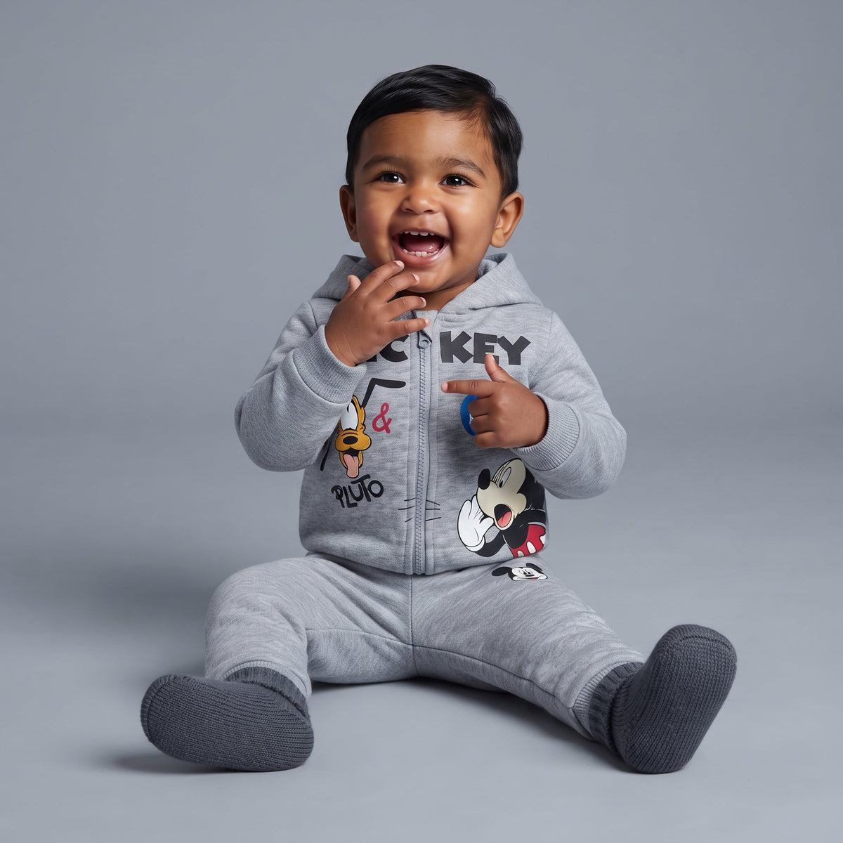 Bebé sonriente vistiendo conjunto EmpiBy de dos piezas gris con estampado de Mickey Mouse y Pluto en sudadera y jogger