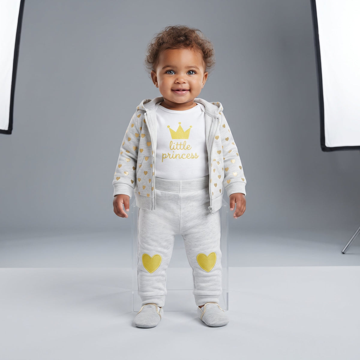Bebé niña sonriente vistiendo conjunto EmpiBy de tres piezas: chaqueta gris con corazones, body blanco y leggings.
