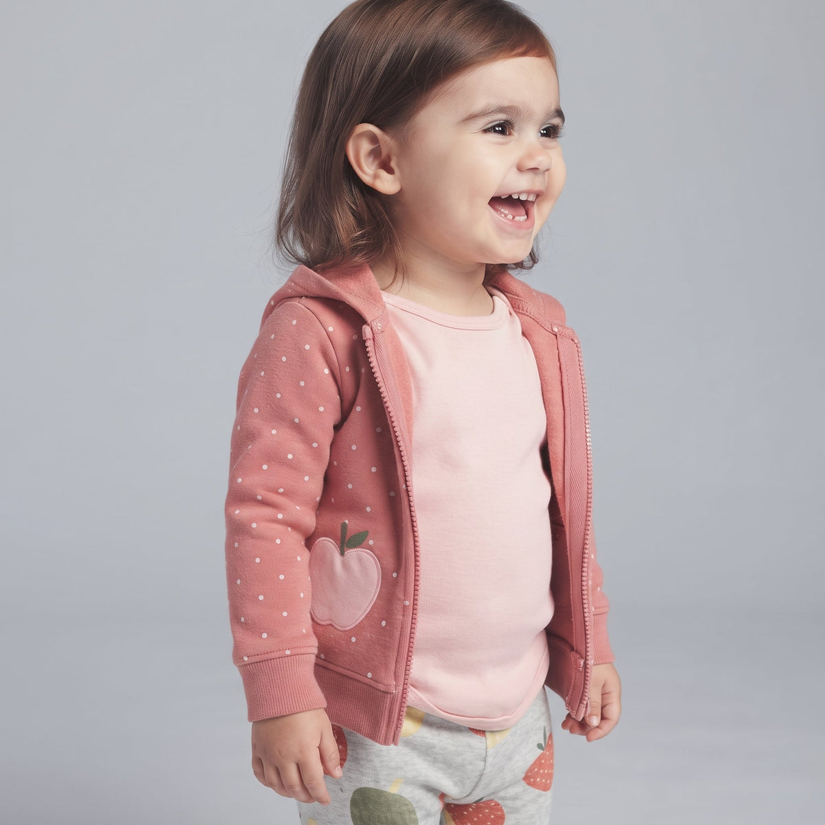 Niña sonriente con conjunto EmpiBy de chaqueta rosa con lunares, body marfil y pantalón estampado de manzanas.