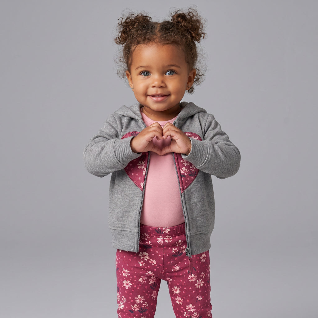 Niña pequeña con conjunto EmpiBy de 3 piezas: chaqueta gris con corazón, body rosa y legging floral frambuesa.