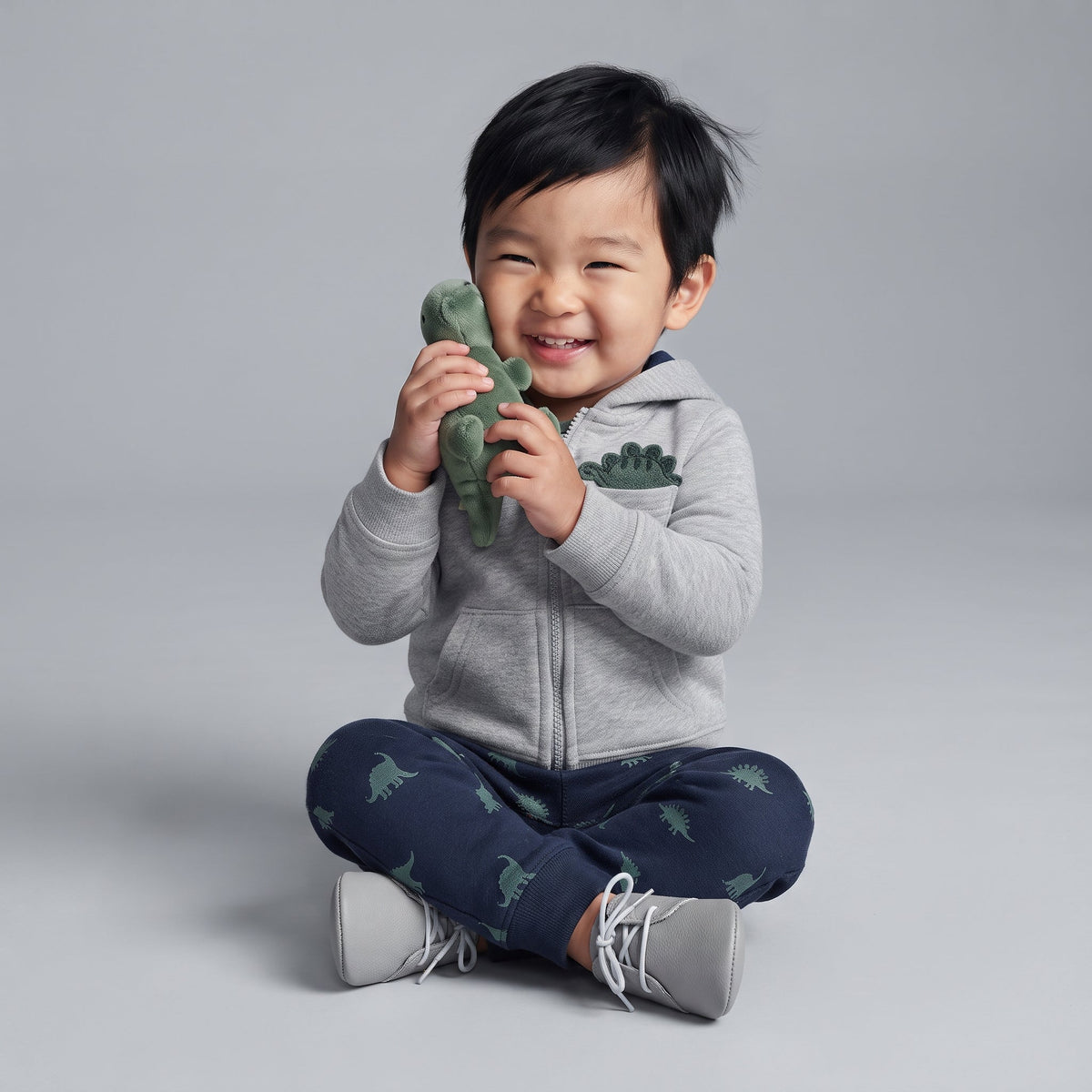 Bebé niño sonriente con conjunto EmpiBy de 3 piezas: chaqueta gris con dinosaurio, body a rayas y pantalón estampado.
