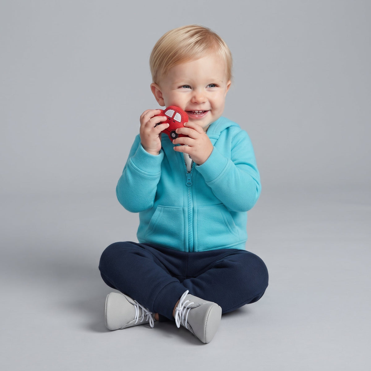 Bebé sonriente con conjunto EmpiBy de chaqueta turquesa con capucha, pantalón azul y coche de juguete rojo