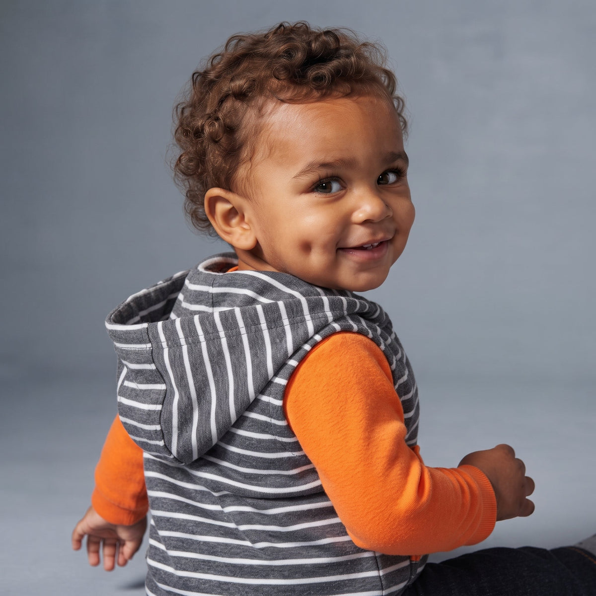 Bebé sonriente con conjunto EmpiBy de chaleco a rayas, body naranja y pantalón gris, ideal para un look moderno.