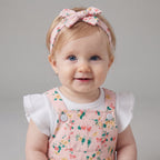Bebé sonriente con conjunto EmpiBy de peto corto rosa con flores, camiseta blanca con volantes y diadema con lazo