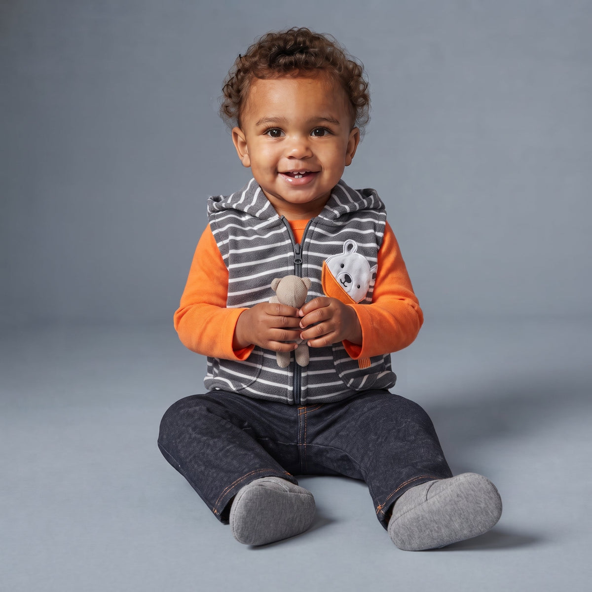 Bebé sonriente con conjunto EmpiBy de 3 piezas: chaleco a rayas, body naranja y pantalón gris oscuro.