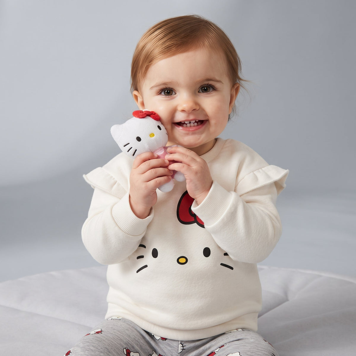 Bebé sonriente con conjunto EmpiBy de sudadera marfil con volantes y legging gris estampado de Hello Kitty