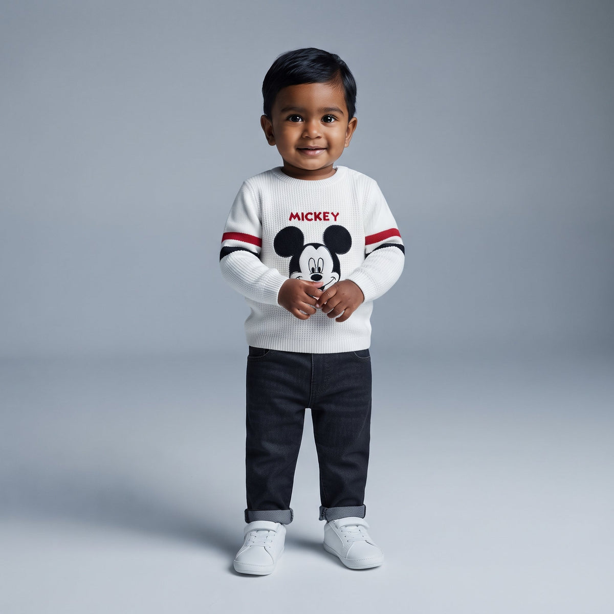 Niño pequeño con conjunto EmpiBy de suéter blanco tejido de Mickey Mouse y pantalón de mezclilla oscuro