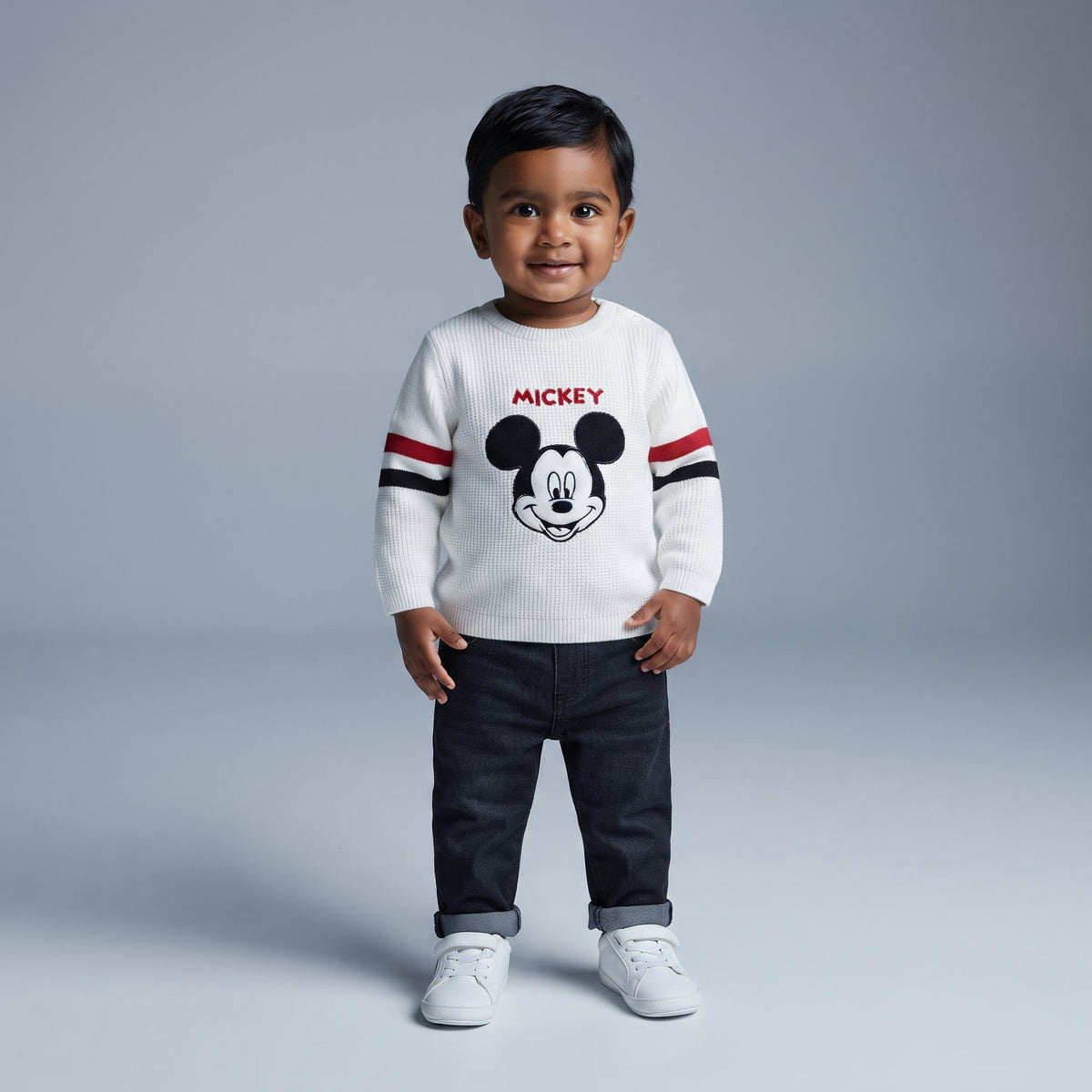 Niño pequeño con suéter blanco tejido de Mickey Mouse y pantalón de mezclilla oscuro de la marca EmpiBy