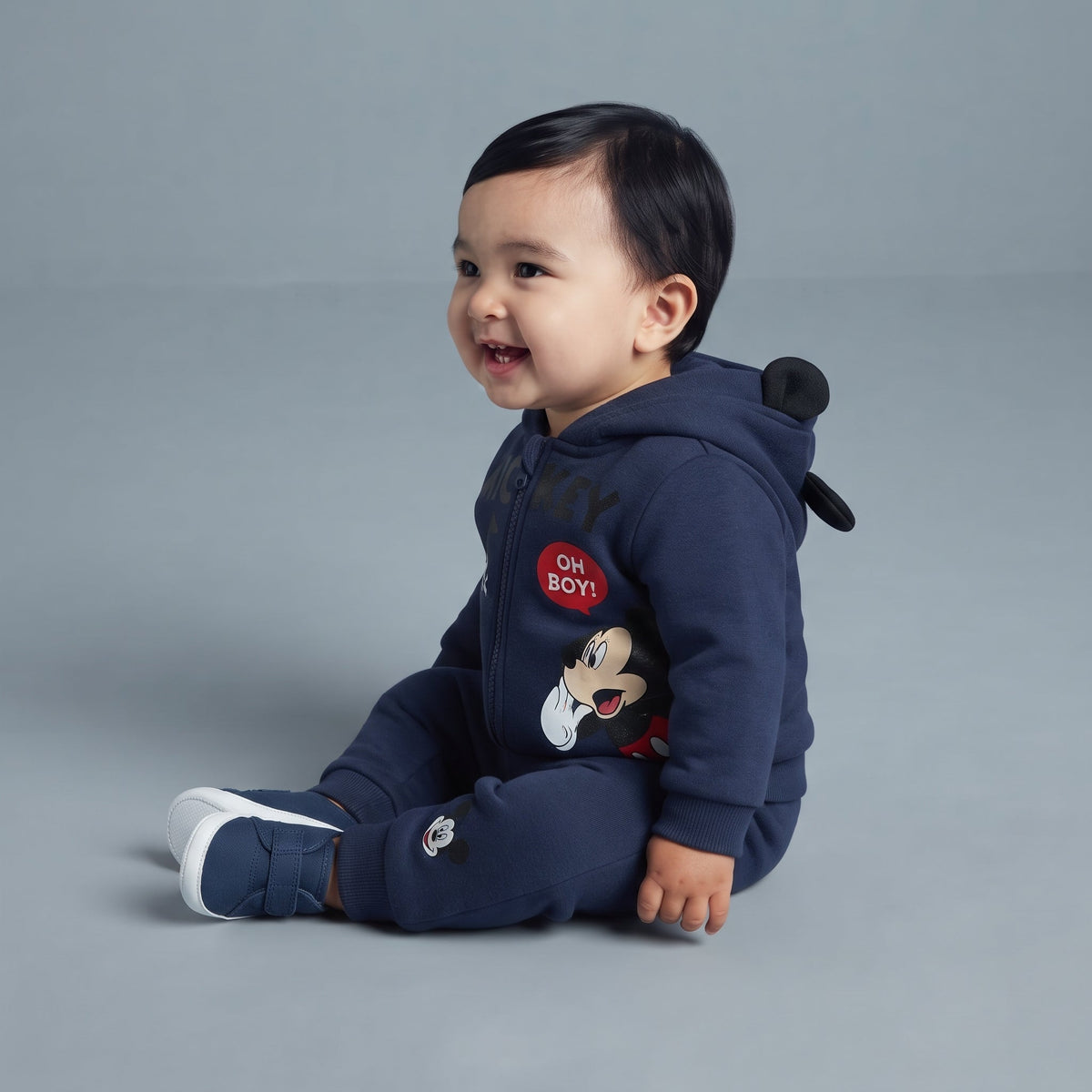 Bebé sonriente vistiendo conjunto EmpiBy de dos piezas azul marino con capucha de orejas de Mickey Mouse y joggers.