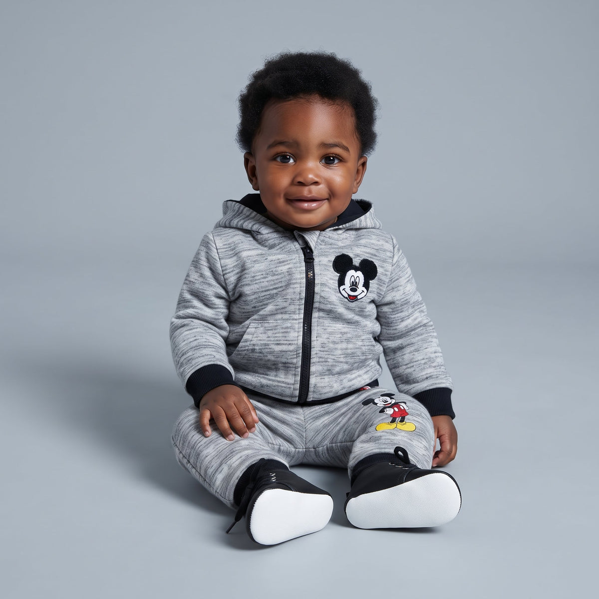 Bebé con conjunto EmpiBy de Mickey Mouse gris melange, sudadera con cremallera, capucha con orejas y pantalón jogger.