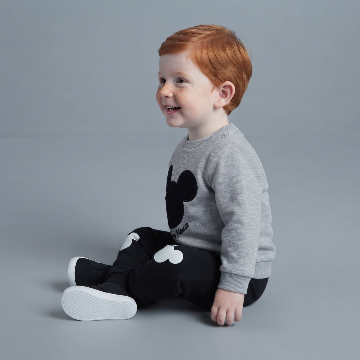 Niño pequeño sonriendo con conjunto EmpiBy de sudadera gris de Mickey Mouse y pantalón jogger negro con estampados.