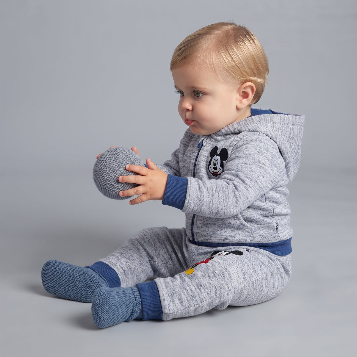 Bebé sentado con conjunto EmpiBy de sudadera gris con capucha y jogger de Mickey Mouse con detalles en azul