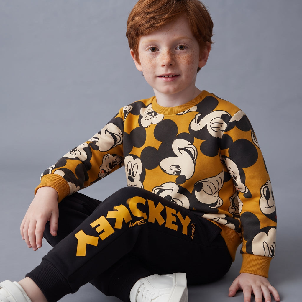 Niño sentado vistiendo conjunto EmpiBy de dos piezas con sudadera y jogger de Mickey Mouse en tonos mostaza y negro