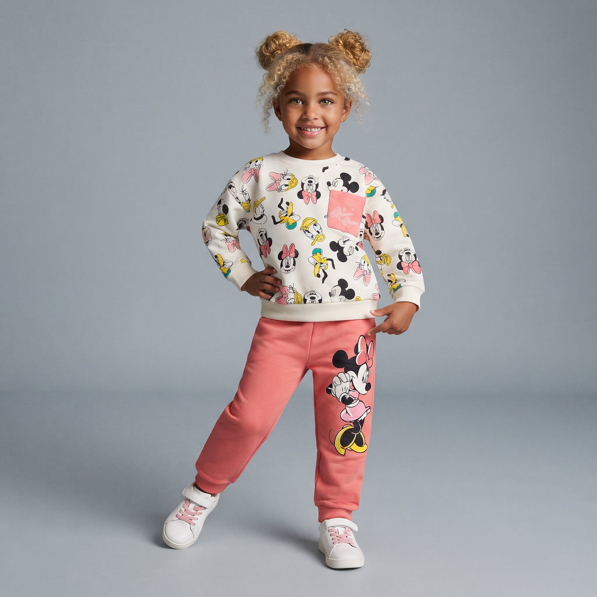 Niña sonriente con conjunto EmpiBy de dos piezas de Minnie Mouse, sudadera estampada y pantalón jogger rosa salmón.
