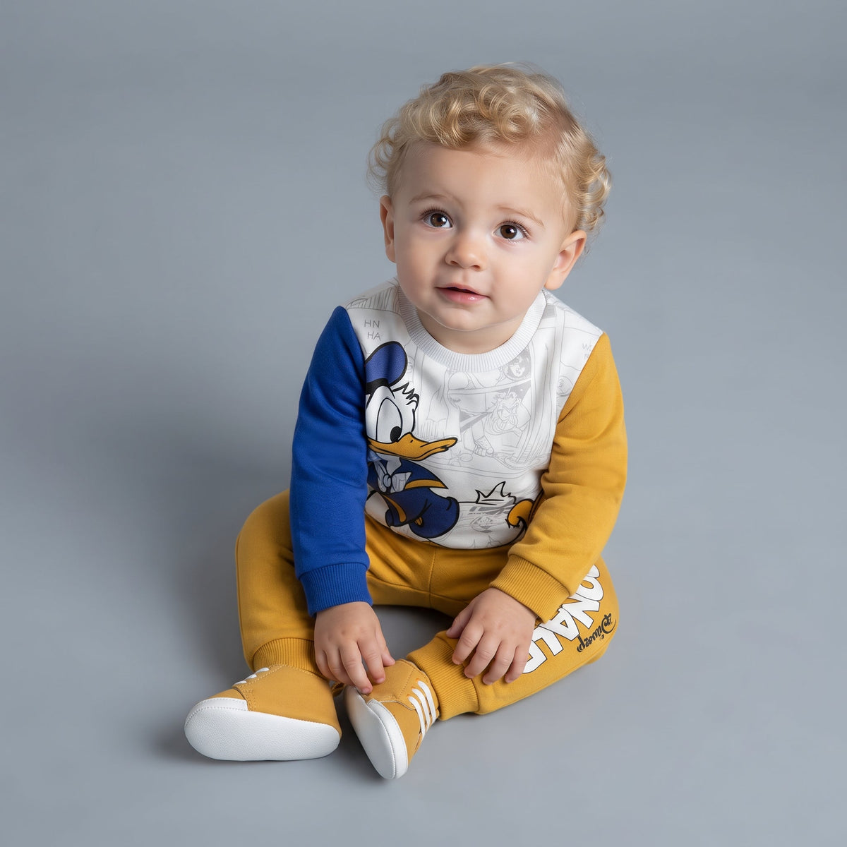 Bebé con conjunto EmpiBy de dos piezas del Pato Donald, sudadera mostaza y azul con jogger a juego en fondo gris.