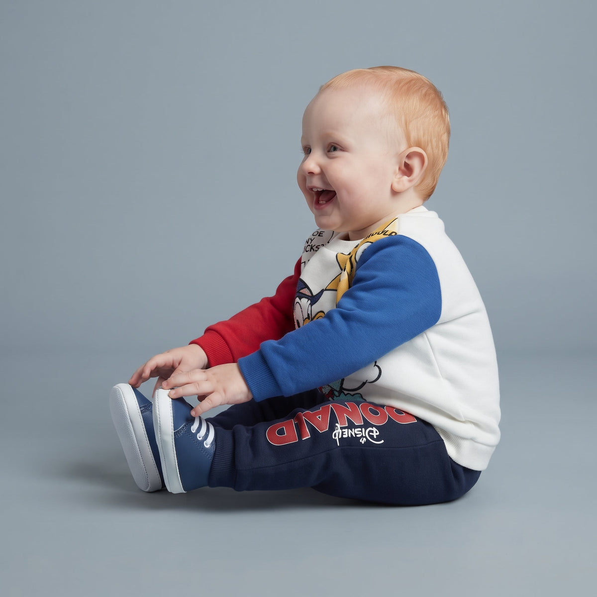 Bebé sonriente vistiendo conjunto EmpiBy de dos piezas del Pato Donald con sudadera multicolor y pantalón jogger azul