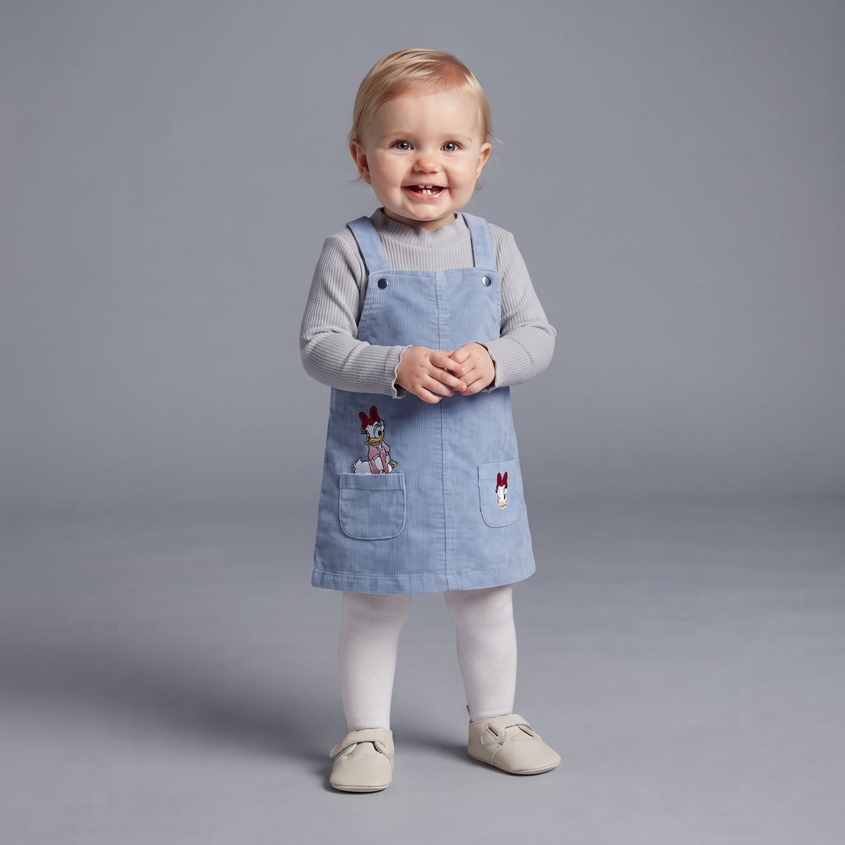 Bebé niña vistiendo conjunto EmpiBy de 3 piezas con vestido de pana azul, camiseta gris y medias blancas de Daisy Duck