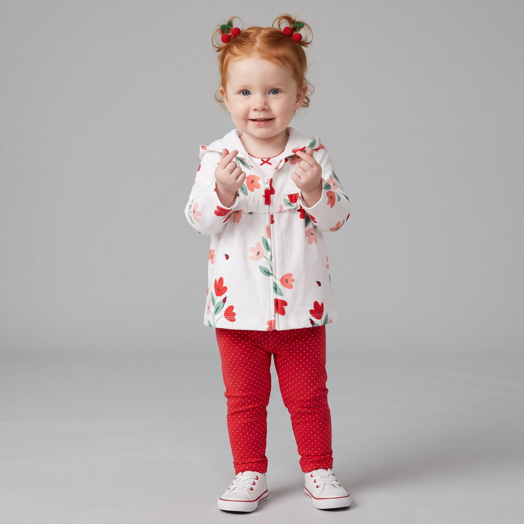 Niña pequeña con conjunto EmpiBy de tres piezas: chaqueta blanca con flores, body y leggings rojos de lunares.