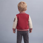 Vista trasera de un niño con conjunto EmpiBy estilo universitario, chaqueta bomber roja y crema y pantalón gris.