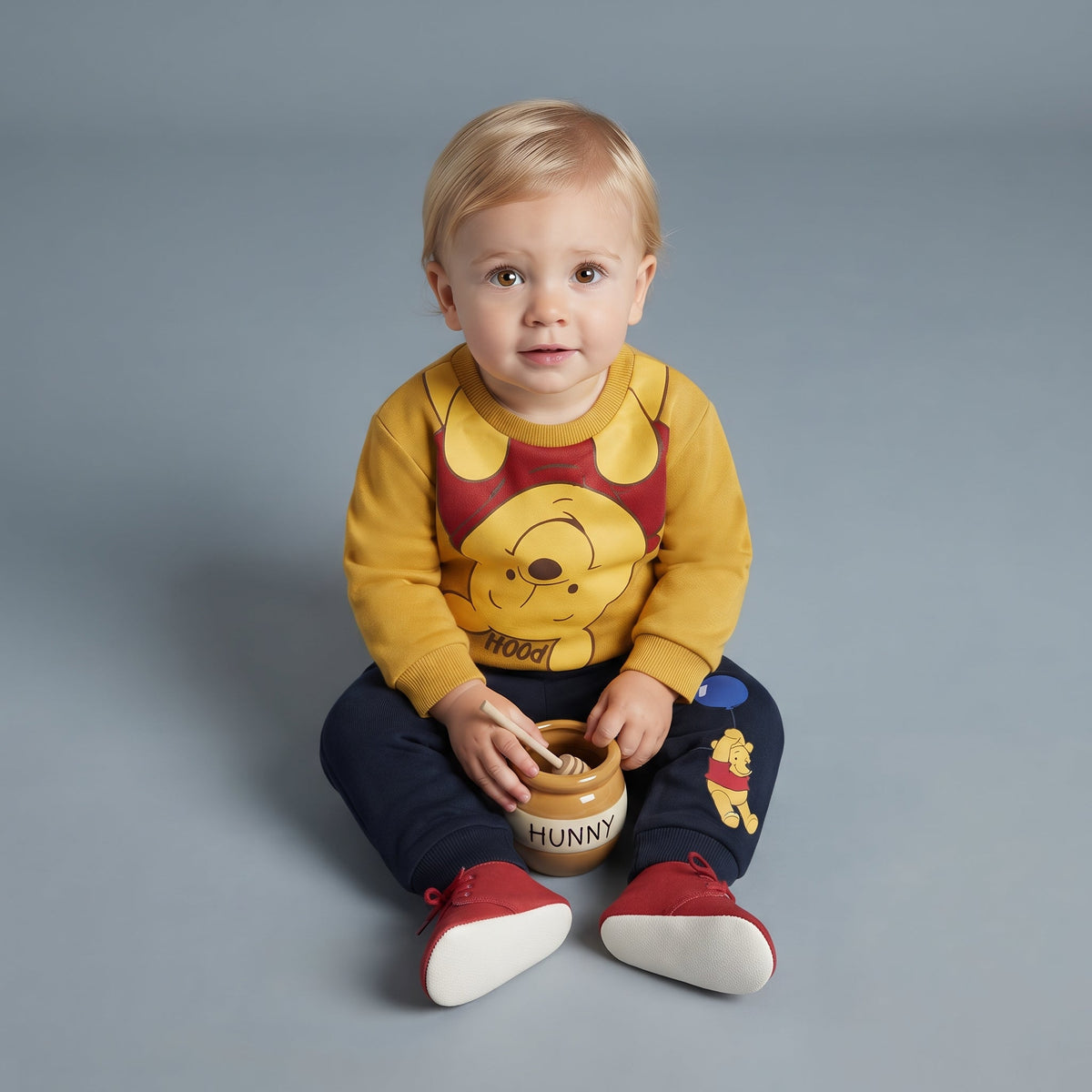 Bebé sentado con conjunto EmpiBy de Winnie Pooh, sudadera amarilla y pantalón jogger azul marino con estampado.